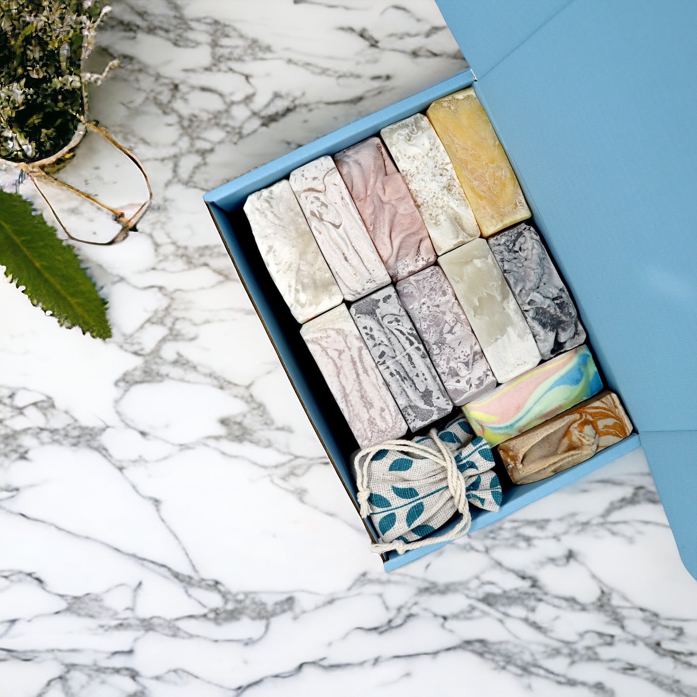Top view of a variety of 12 soap bars inside a blue gift box, and a surprise item in a blue patterned bag with drawstring, displayed on a marble counter top with a plant nearby