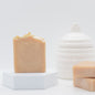 A bar of oat milk and honey soap is displayed on a small white platform next to a stack of 2 bars of soap in front of a white honey jar