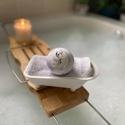 White and purple bath bomb in a bath tub-shaped holder placed on a bath caddy over a bubble bath