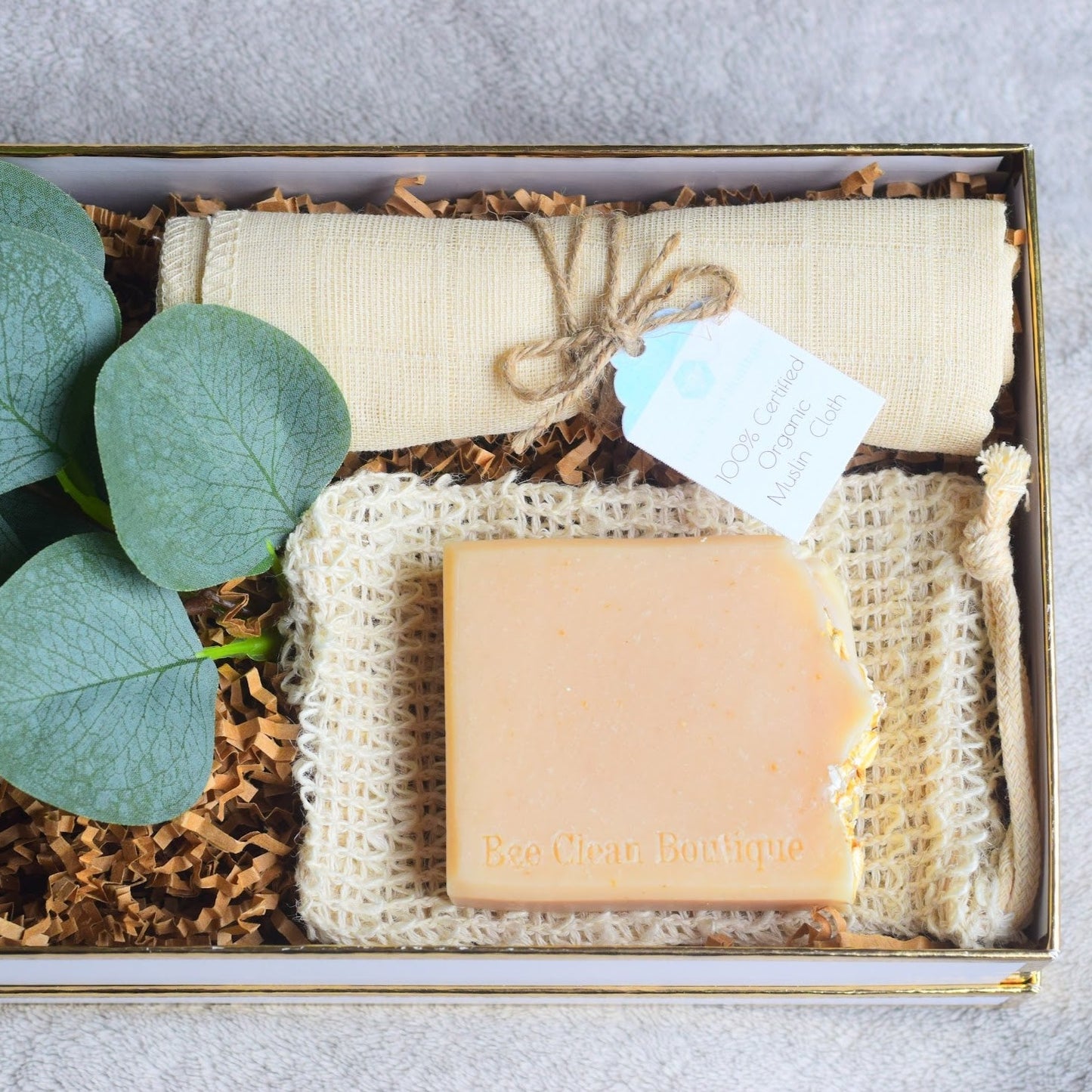 An open gift box showing a bar of artisan soap over a sisal pouch, with a muslin cloth and greenery over kraft crinkle paper