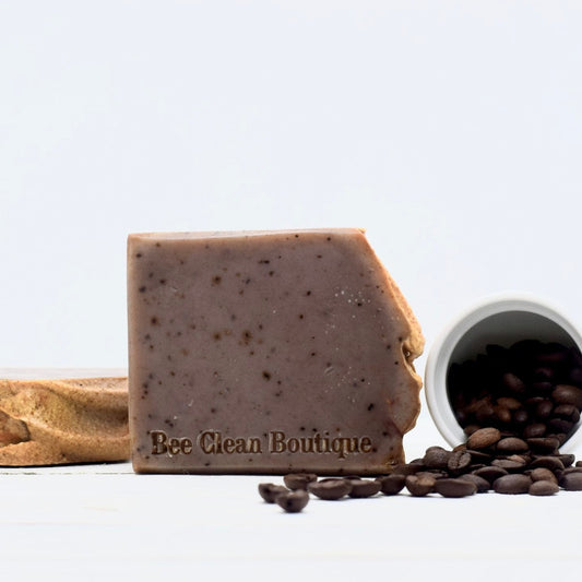 A bar of coffee shop soap featured in front of another bar lying flat and a jar of coffee beans pouring out on a white surface