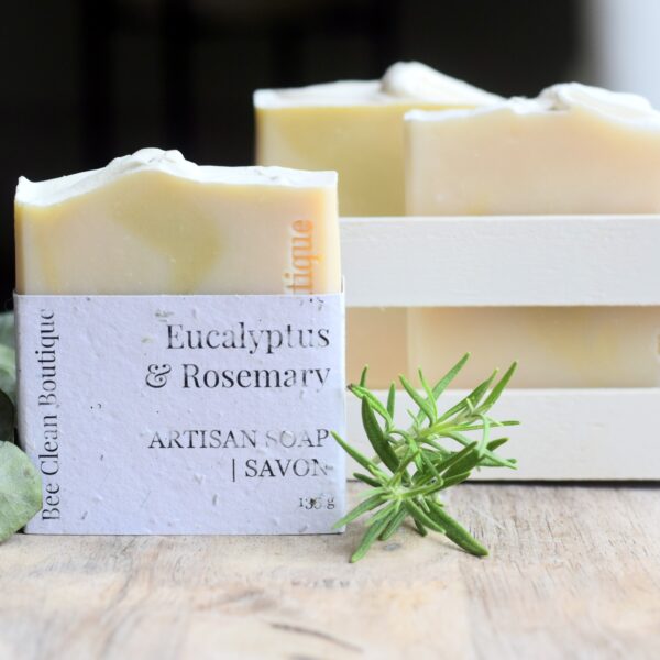 A eucalyptus and rosemary bar soap displayed in front of a small crate of bar soaps; a sprig of rosemary is next to the featured soap