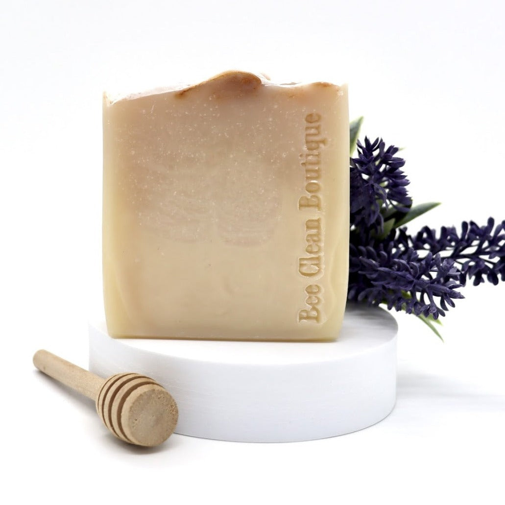 A bar of handmade lavender honey soap is displayed on a white platform, surrounded by lavender and a wooden honey dipper