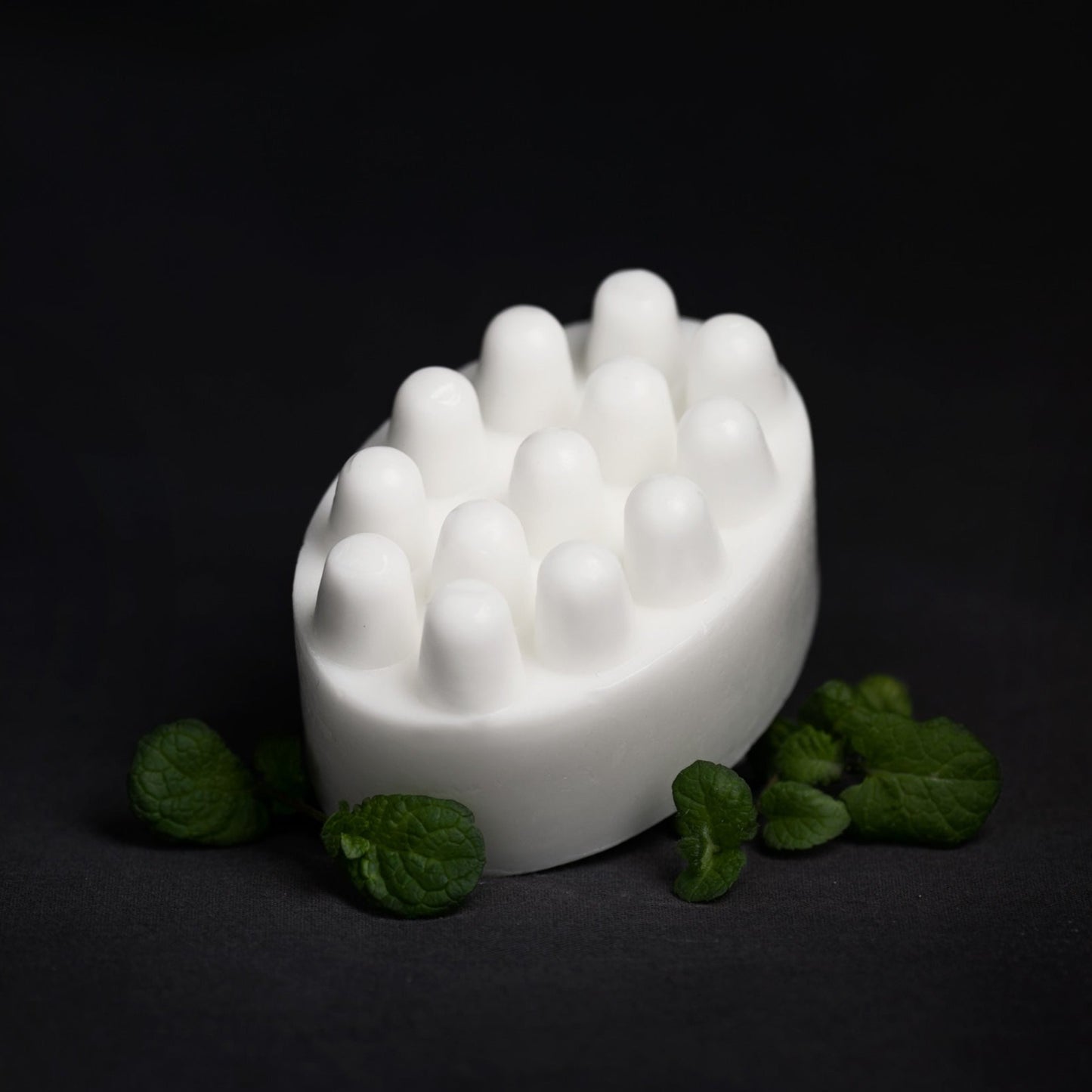 a white bar of soap with massage bumps, surrounded by mint leaves on a black surface