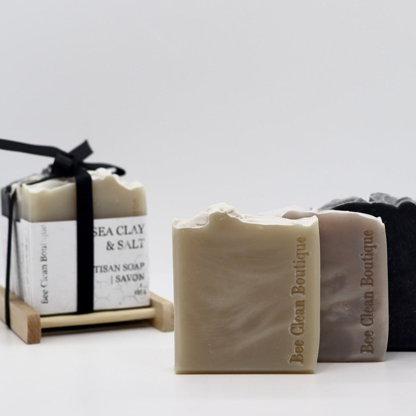 3 bars of loose bar soaps displayed in front of a set of soaps tied together with black ribbon on a wooden soap dish