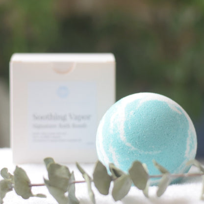 a blue and white bath bomb displayed in front of a white paper box and a stem of eucalyptus in front